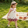 A young girl wearing a cream summer pinafore dress with strawberry and bunny print, standing in a grassy field.