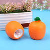 Juguete de zanahoria exprimible naranja con tapa verde junto a una maceta sobre una superficie azul.