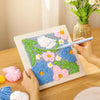 Hænder samler et blomster punch needle broderi sæt med farverige garnblomster på en træplade.