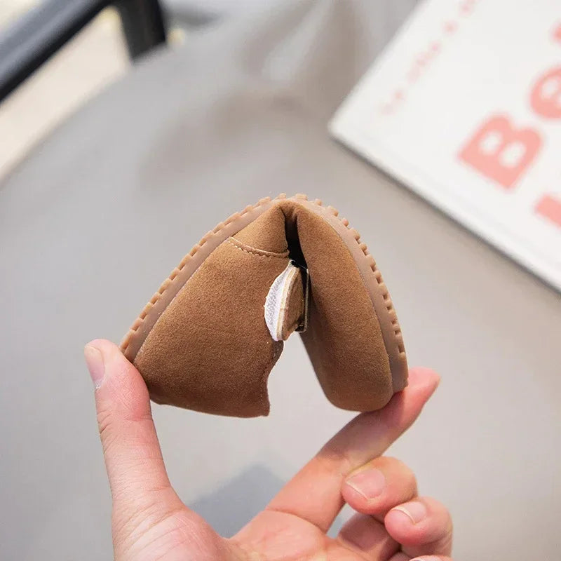 A hand holds a brown retro non-slip kids' moccasin shoe with an easy hook-and-loop closure.