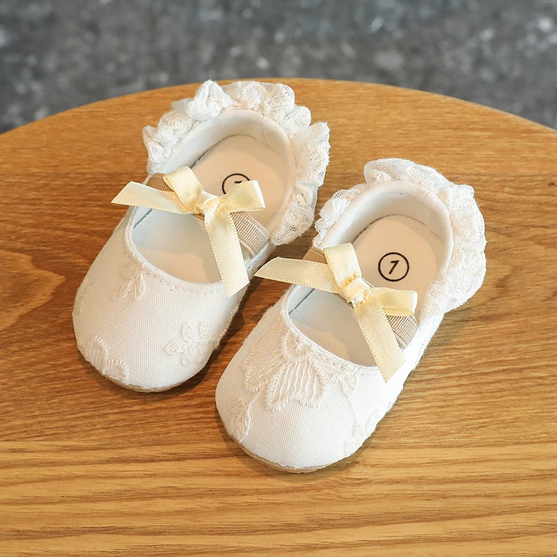 Pair of white baby first walker shoes with lace trim and a yellow ribbon on a wooden surface.