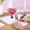 A hand is writing on a pink card featuring a 3D pop-up love tree and a silhouette of a couple.