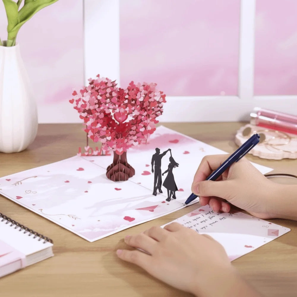 A hand is writing on a pink card featuring a 3D pop-up love tree and a silhouette of a couple.