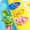 A hand holds a green kids shark clog sandal with a red accent and an yellow kids shark clog sandal.