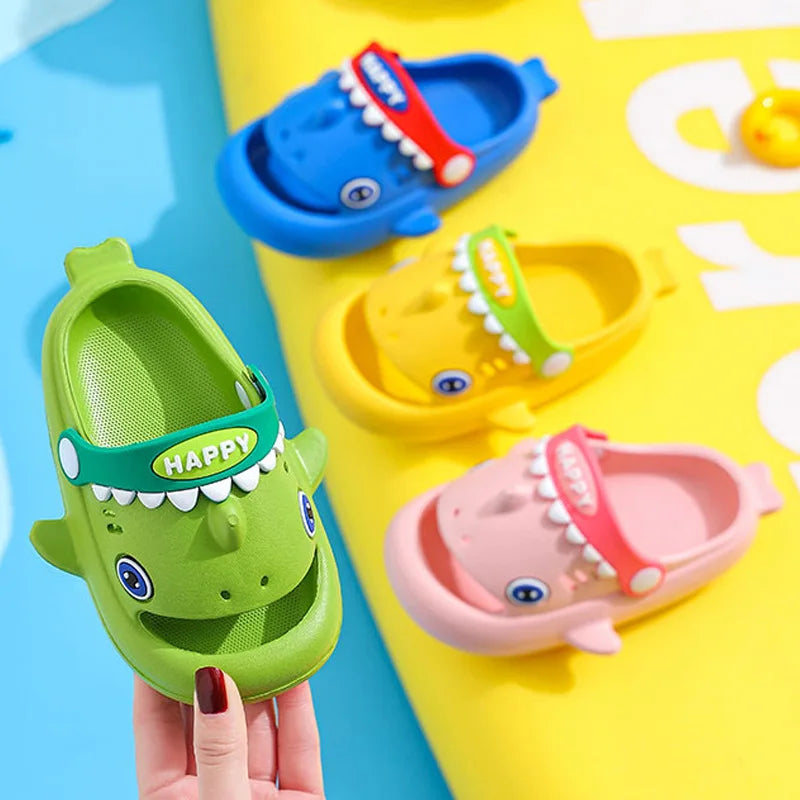 A hand holds a green kids shark clog sandal with a red accent and an yellow kids shark clog sandal.
