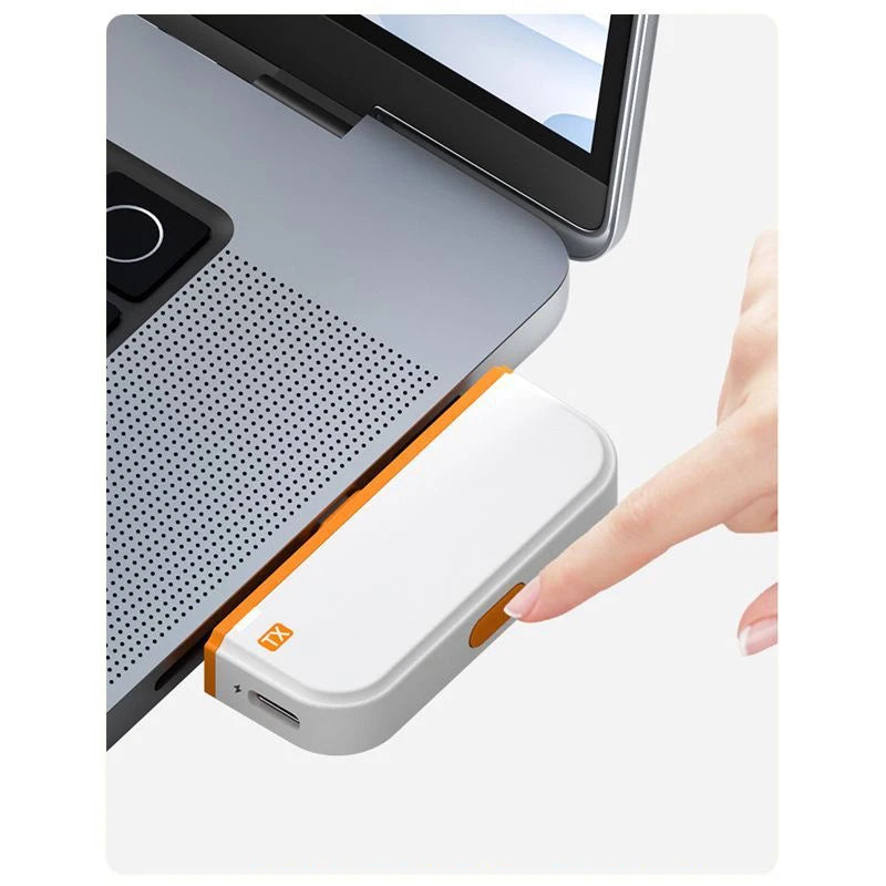 Käsi pitää valkoista USB-C langatonta HDMI-lähetintä oranssilla korostuksella, hopeisen kannettavan tietokoneen vieressä.