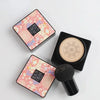 Paquete cuadrado de crema de belleza rosa con aplicador de cabeza de hongo negro sobre superficie blanca.