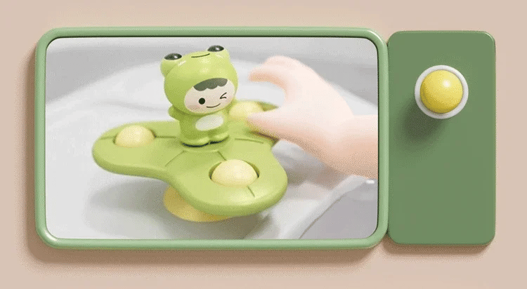 A green suction cup spinner bath toy shaped like a cute animal is being held by a hand, with a yellow ball nearby.