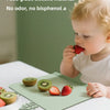 A baby is eating fruit from a green silicone feeding placemat on a white table.