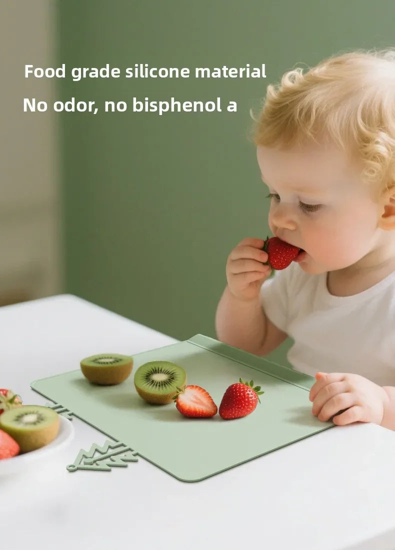 A baby is eating fruit from a green silicone feeding placemat on a white table.
