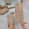 A pair of tan suede knee-high boots with chunky heels and a metal buckle are displayed on a light floor.