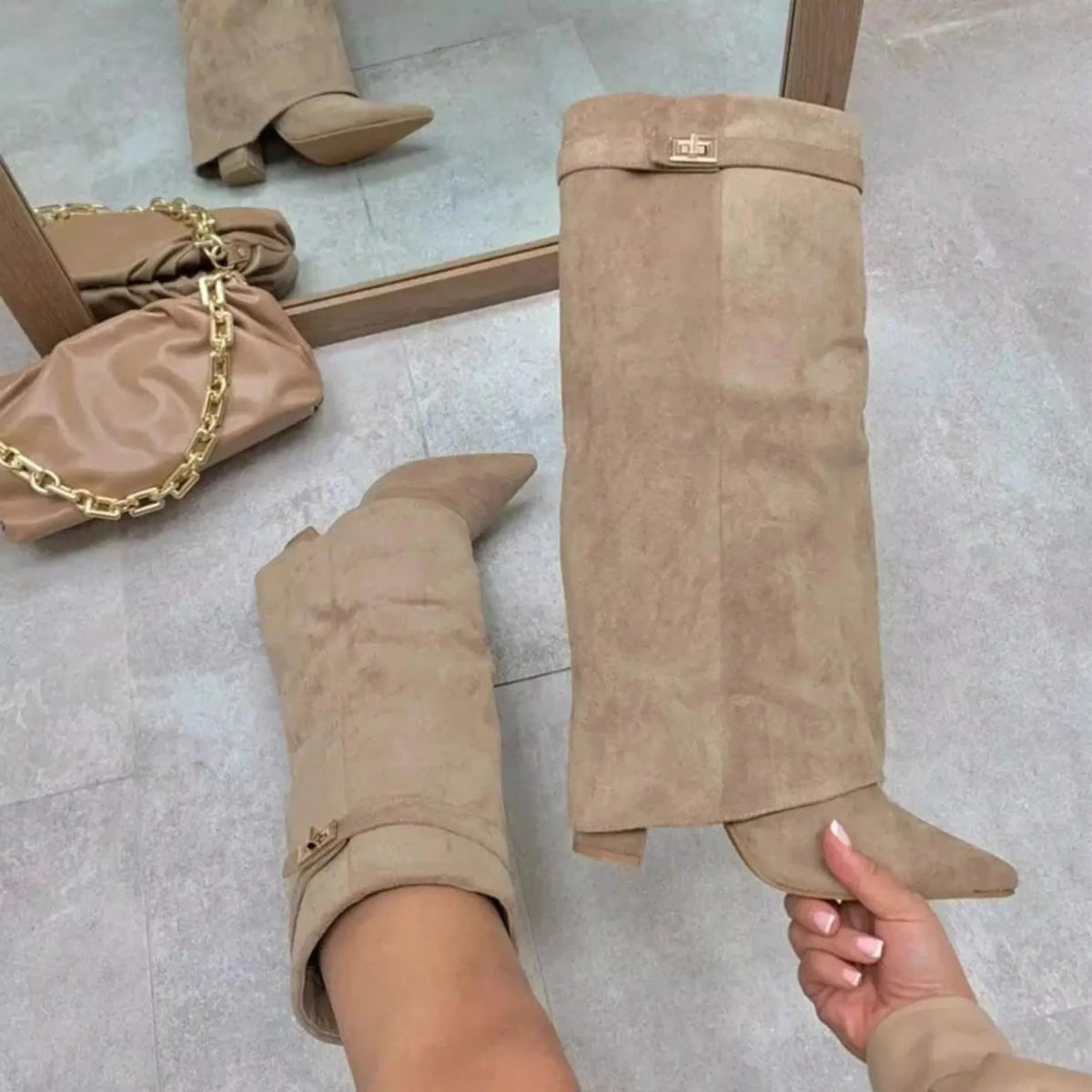 A pair of tan suede knee-high boots with chunky heels and a metal buckle are displayed on a light floor.