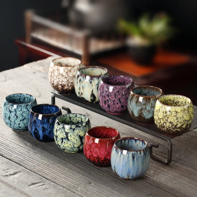 Juego de tazas de té de cerámica con esmalte tenmoku en tonos verde, azul y morado sobre una bandeja oscura.