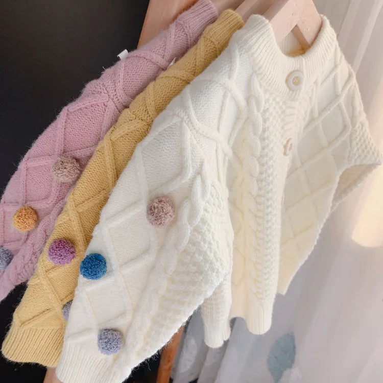 A white knitted cardigan sweater with colorful pom-pom trim displayed on a wooden hanger.