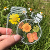 A hand holds a clear glass jar containing three dried flowers used as bookmarks in a natural outdoor setting.