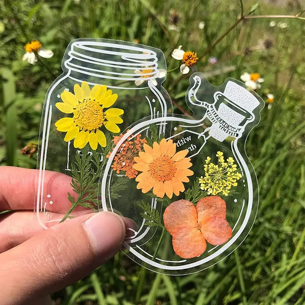 A hand holds a clear glass jar containing three dried flowers used as bookmarks in a natural outdoor setting.