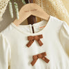 A white long-sleeve knit dress with brown fabric bows, displayed on a wooden hanger.