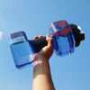 Una mano sostiene una botella de agua deportiva azul con forma de pesa y tapa negra, contra un cielo azul claro.