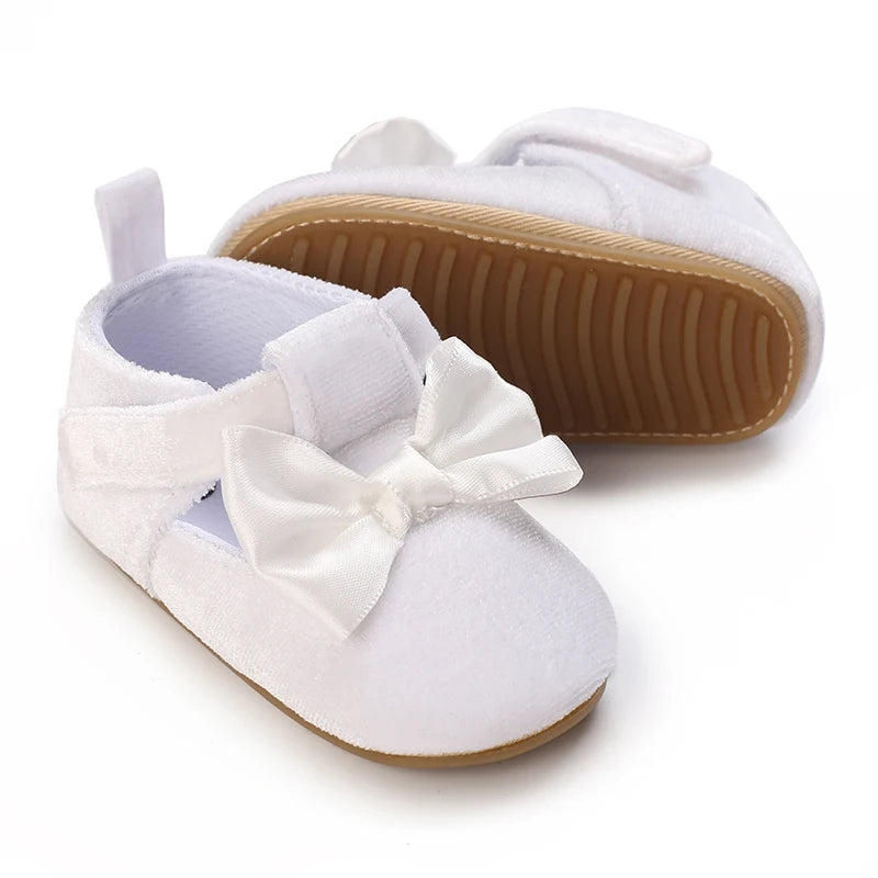 A pair of white soft non-slip baby first walker shoes with a bow detail and tan sole.
