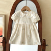 A cream-colored girls summer dress with floral embroidery displayed on a wooden hanger inside a large mirror.