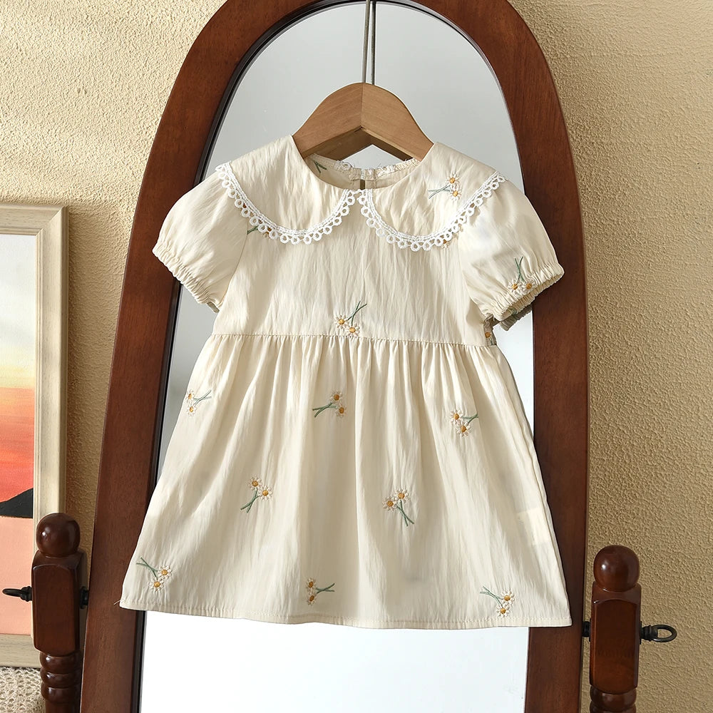 A cream-colored girls summer dress with floral embroidery displayed on a wooden hanger inside a large mirror.