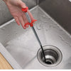 A hand is using a flexible drain snake with a red handle to clear a clog in a stainless steel kitchen sink.