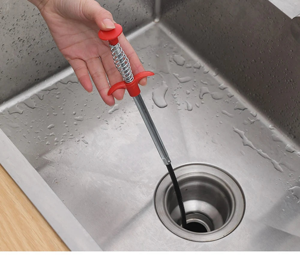 A hand is using a flexible drain snake with a red handle to clear a clog in a stainless steel kitchen sink.
