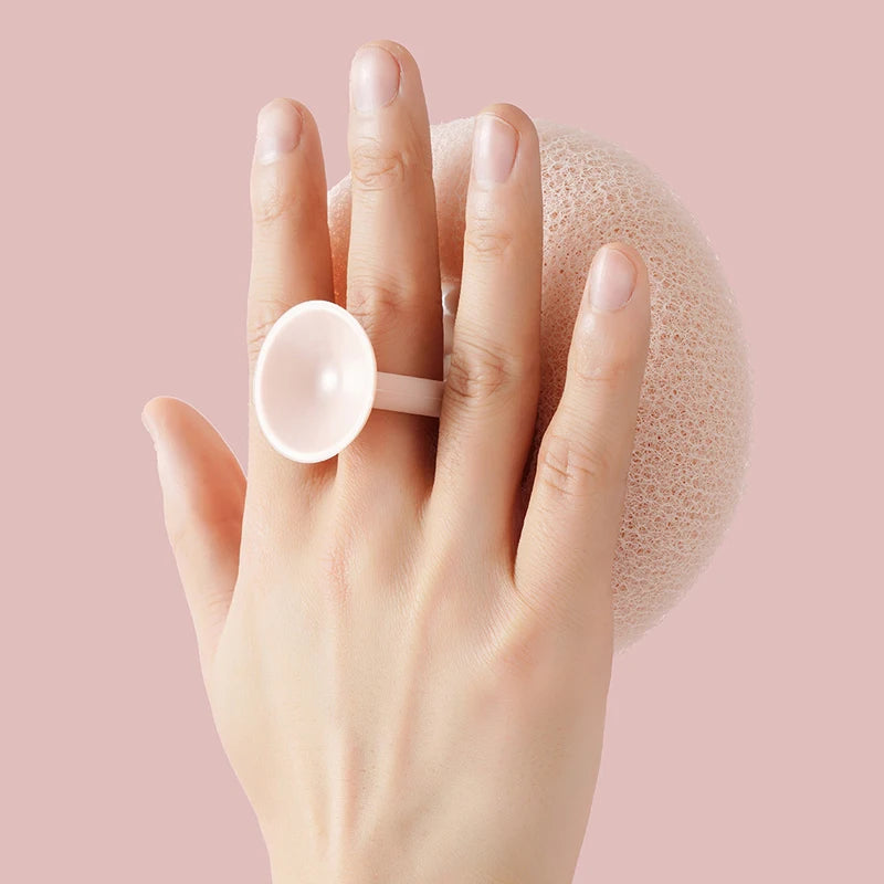 A hand holds a pink bath sponge with a white suction cup on a light pink textured surface.