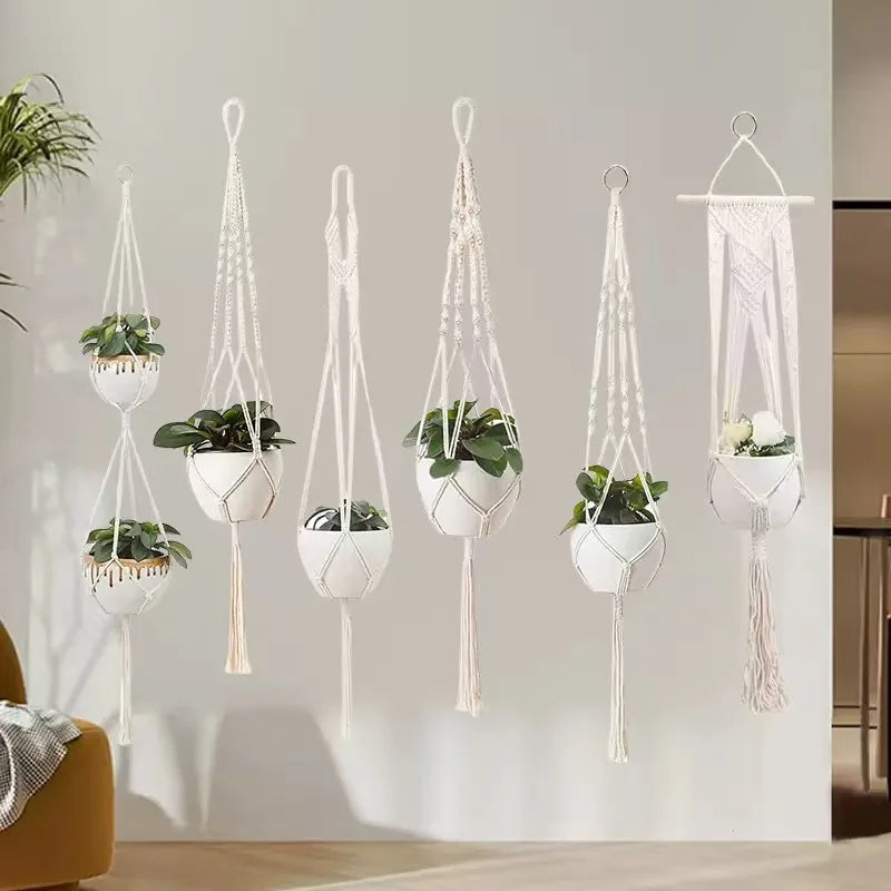 En samling hvite makrame-plantekasser med ulike grønne planter hengende mot en lys grå vegg.