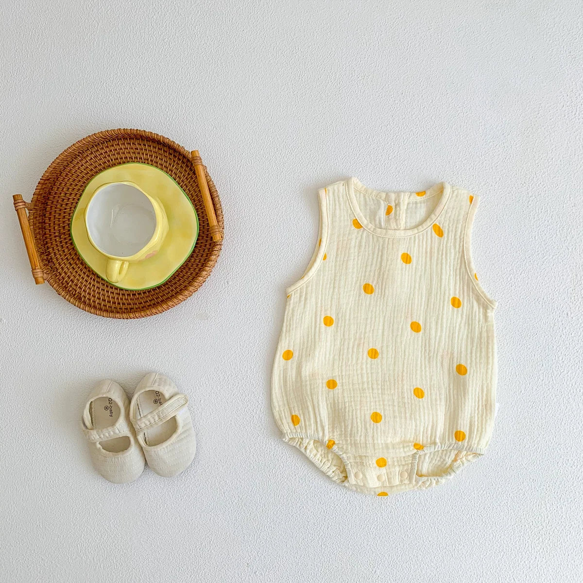 A cream-colored baby cotton sleeveless jumpsuit bodysuit set with small orange polka dots, next to a woven tray and a pair of