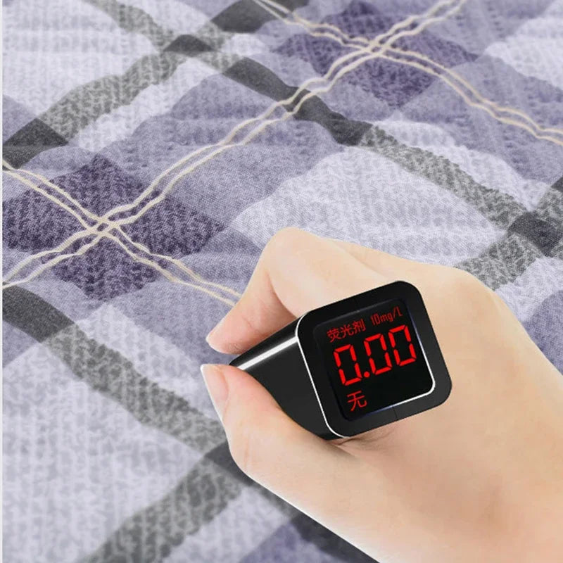 A hand holds a black digital blood pressure monitor displaying 0.00, resting on a plaid fabric background.