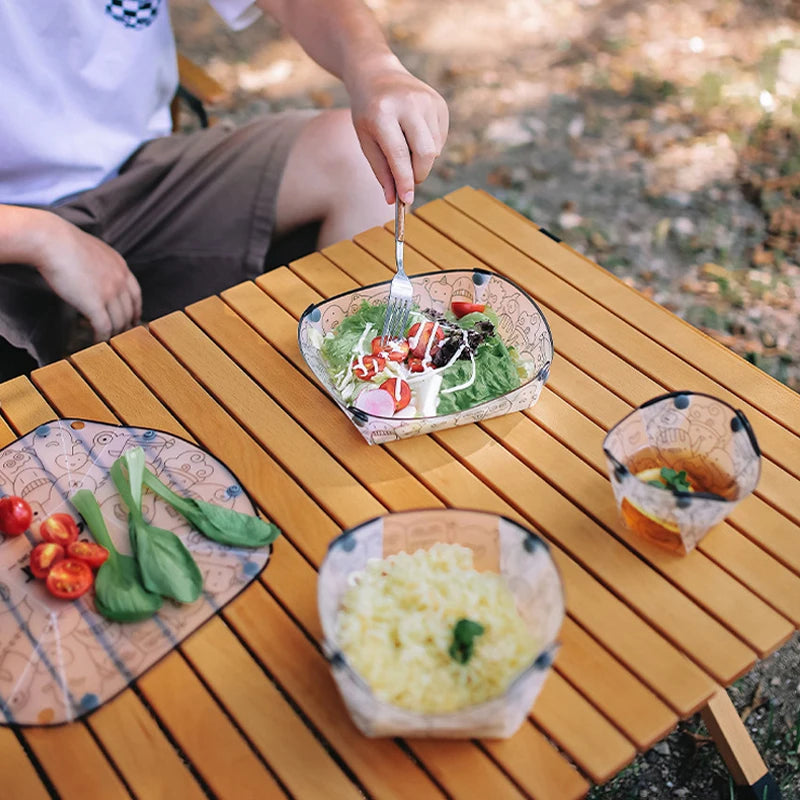 A wooden table set for outdoor dining featuring a bowl of rice, a plate with food, and a cup.