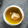 En keramikk dessertskål fylt med en gyllengul suppe eller buljong, pyntet med et stykke toast og en kvist grønn urt.