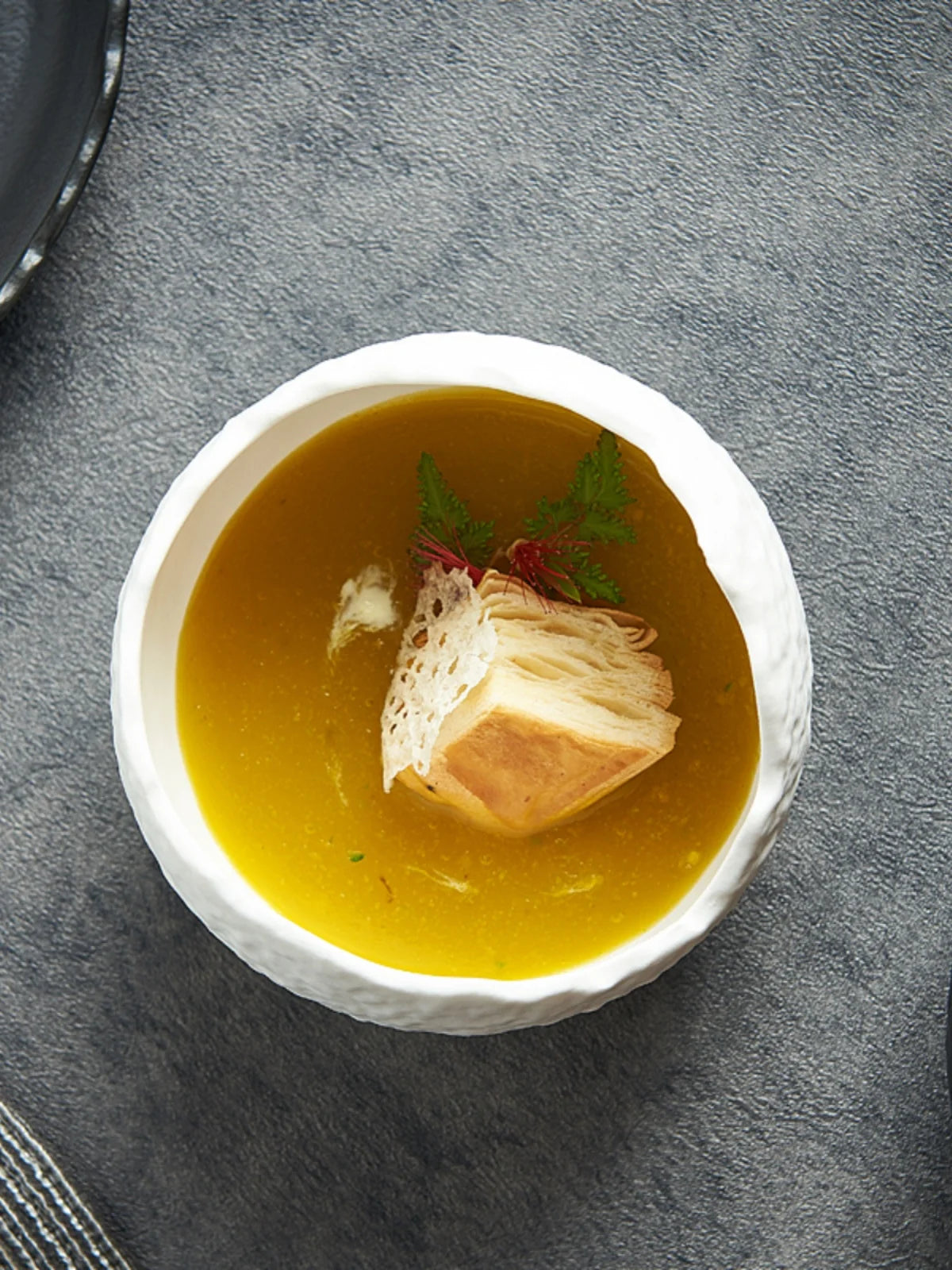 En keramikk dessertskål fylt med en gyllengul suppe eller buljong, pyntet med et stykke toast og en kvist grønn urt.