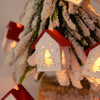 Ein hölzerner Weihnachtshaus-Anhänger mit beleuchtetem Tannenbaum-Ausschnitt hängt an einem verschneiten immergrünen Zweig.