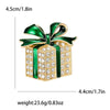 Broche de caja de regalo navideña en esmalte dorado con pedrería y cinta verde.