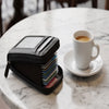 A black compact zipper card holder is resting on a white marble table next to a white coffee cup.