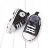 A pair of black and white canvas baby sneakers with a soft non-slip sole resting on a white wooden surface.
