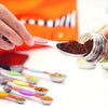 A hand uses a stainless steel measuring spoon to scoop dark powder into a glass jar, with other spoons visible nearby.