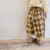 A child wearing beige top and olive green diamond patterned harem pants with light brown shoes.