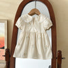 A cream-colored girls summer dress with floral embroidery and a Peter Pan collar hanging in a wooden arched mirror.