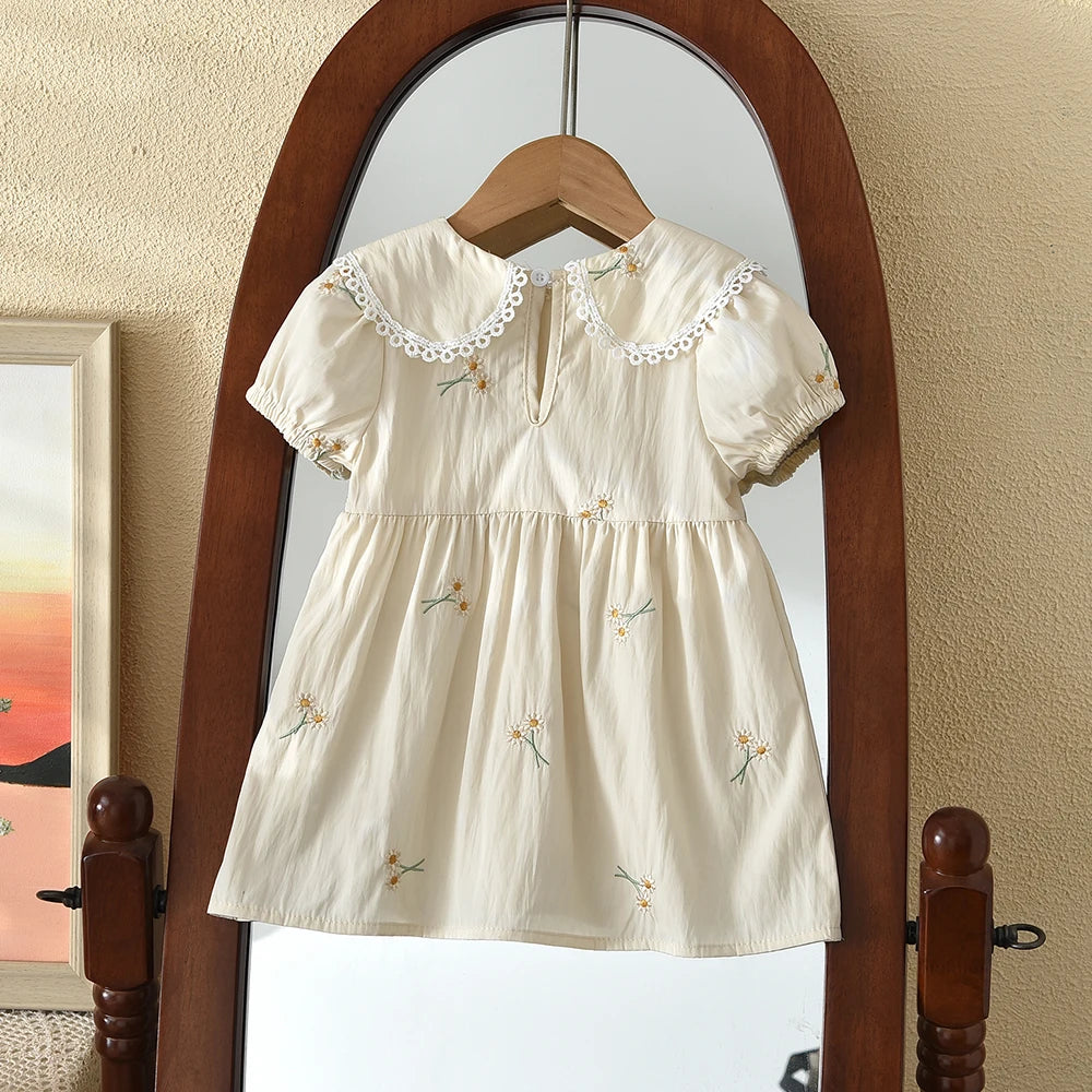 A cream-colored girls summer dress with floral embroidery and a Peter Pan collar hanging in a wooden arched mirror.