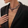 A close-up of a woman wearing a silver and black boho tassel pendant necklace with a beaded cross detail against a black blaz