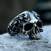 A detailed gothic carved skull ring made of silver-toned stainless steel resting on a gray surface.