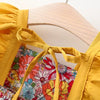 A yellow dress with a ruffled neckline and tie detail is displayed on a wooden hanger against a floral print fabric.