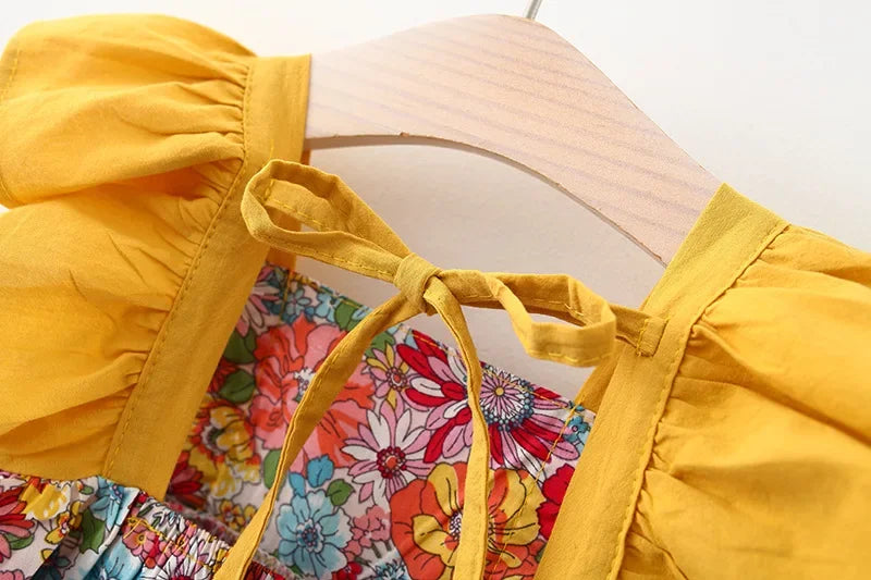 A yellow dress with a ruffled neckline and tie detail is displayed on a wooden hanger against a floral print fabric.
