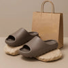A pair of taupe EVA platform slides resting on light-colored stones next to a brown paper shopping bag.