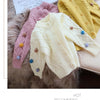 A display of three knitted cardigans in pink, cream, and yellow with pom-pom trim.