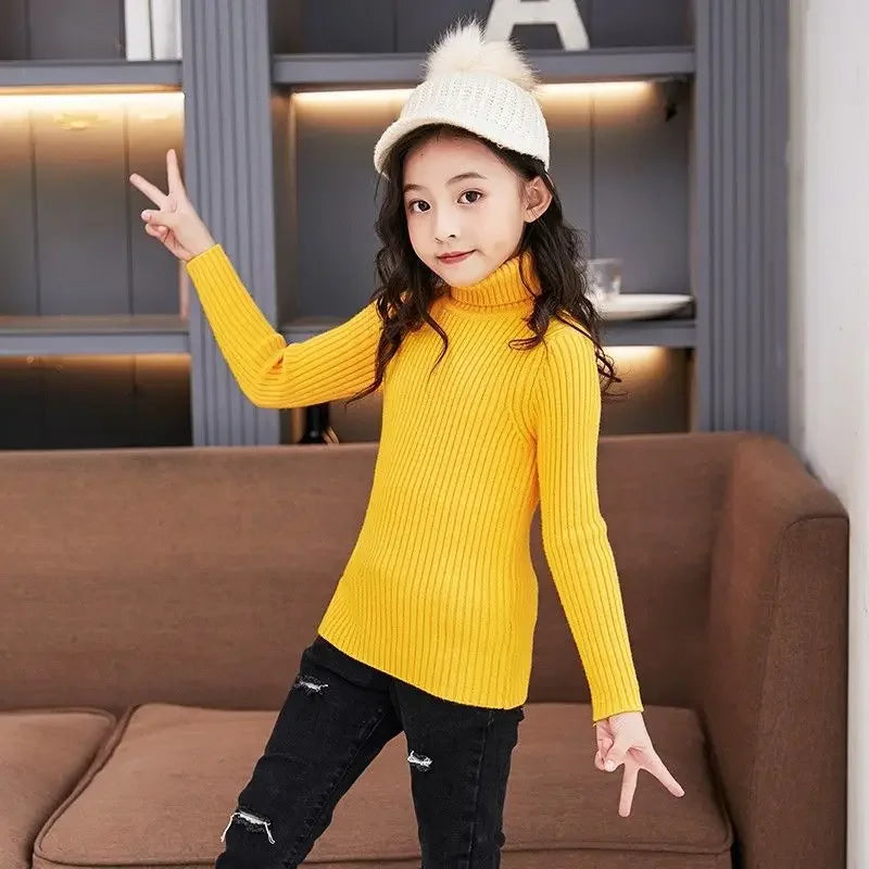 A young girl wearing a yellow knit turtleneck sweater and black jeans posing with her fingers raised.