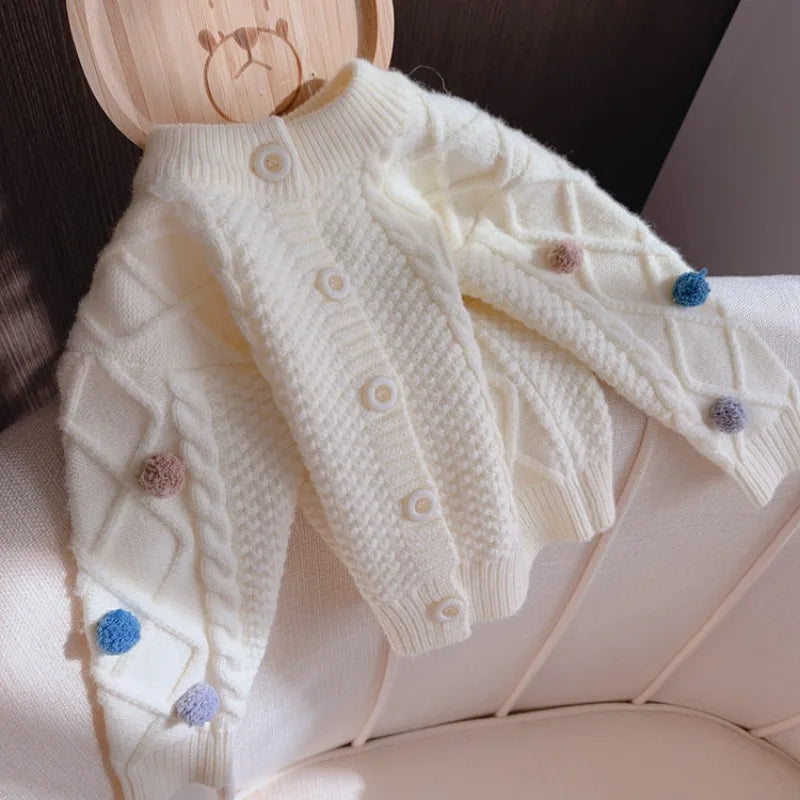A white knitted cardigan sweater for girls featuring pom-pom trim details.